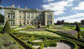 The grand façade of Sledmere House near Bungalow Farm in Buckton, overlooking formal gardens with geometric hedges, colourful flowerbeds and a central statue under bright summer light.
