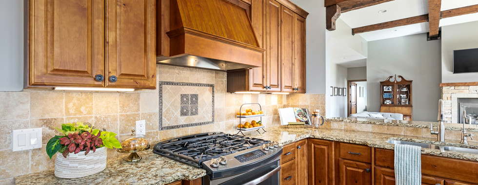Warm, inviting kitchen with natural wood cabinets, granite countertops, and a tiled backsplash centered around a gas range and decorative hood. Exposed ceiling beams add rustic charm.