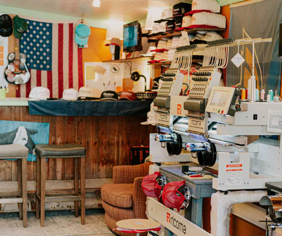 Embroidery machine in a cozy room with wood accents. Hats displayed, an American flag on the wall, and spools of thread add color.