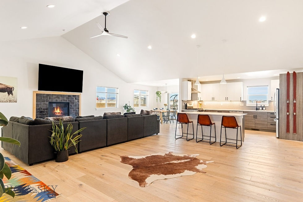 Open living room with fireplace, TV, dark sofa, and colorful rug. Kitchen with island and bar stools. Bright, spacious, modern design.