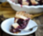 Slice of berry pie on a white plate with a fork, set on a rustic wooden table. The pie has a golden crust and juicy purple filling.