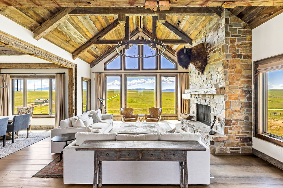 Rustic living room with stone fireplace, mounted bison head, white sofas, and large windows overlooking a green landscape. Earthy tones.