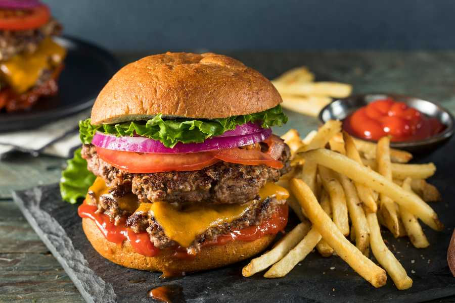 Cheeseburger with lettuce, tomato, onion, and ketchup on a bun, served with fries and a side of ketchup on a dark textured surface.