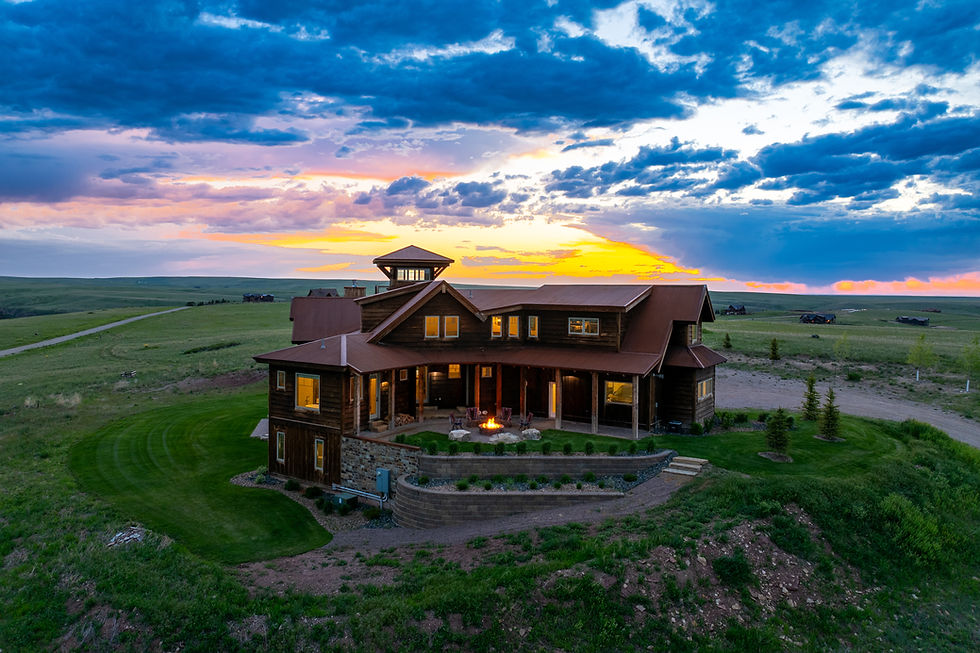 Wooden house with warm glowing windows at dusk, set in a vast grassy field with a dramatic blue sky and rolling hills in the background.