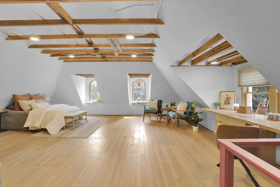 Spacious attic room with exposed beams, a cozy bed, two teal chairs, wooden desk, a slanted roof, and soft lighting from windows.