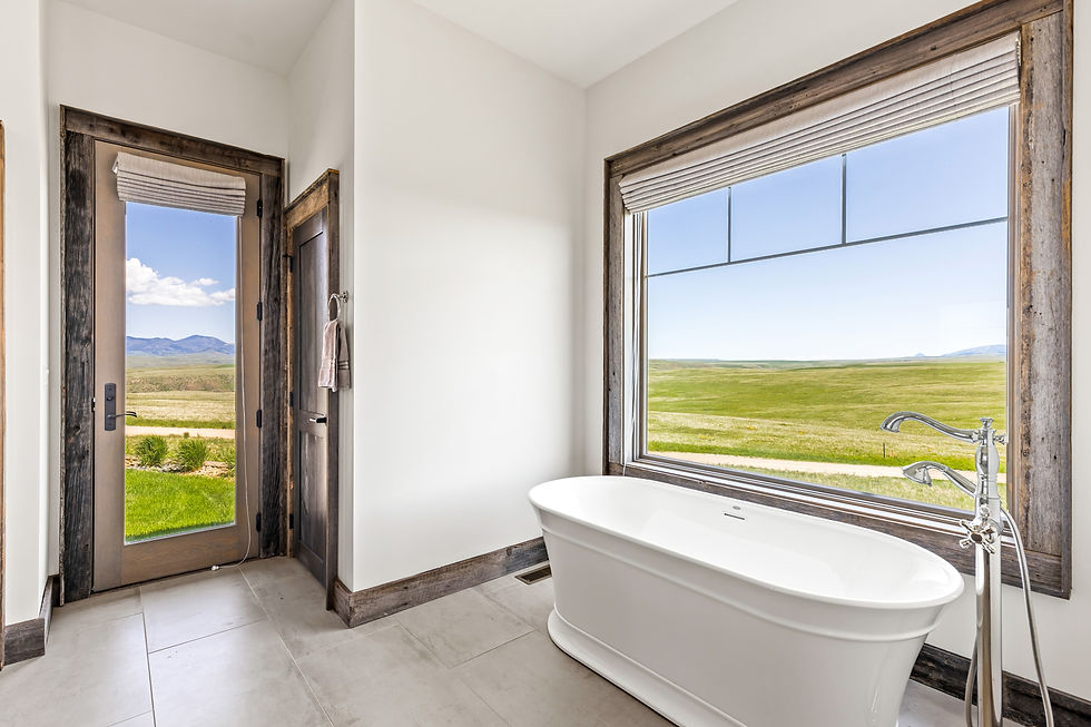 Freestanding bathtub in a bright bathroom with large window showing a vast green field and mountains. Rustic wood frame and beige tiles.