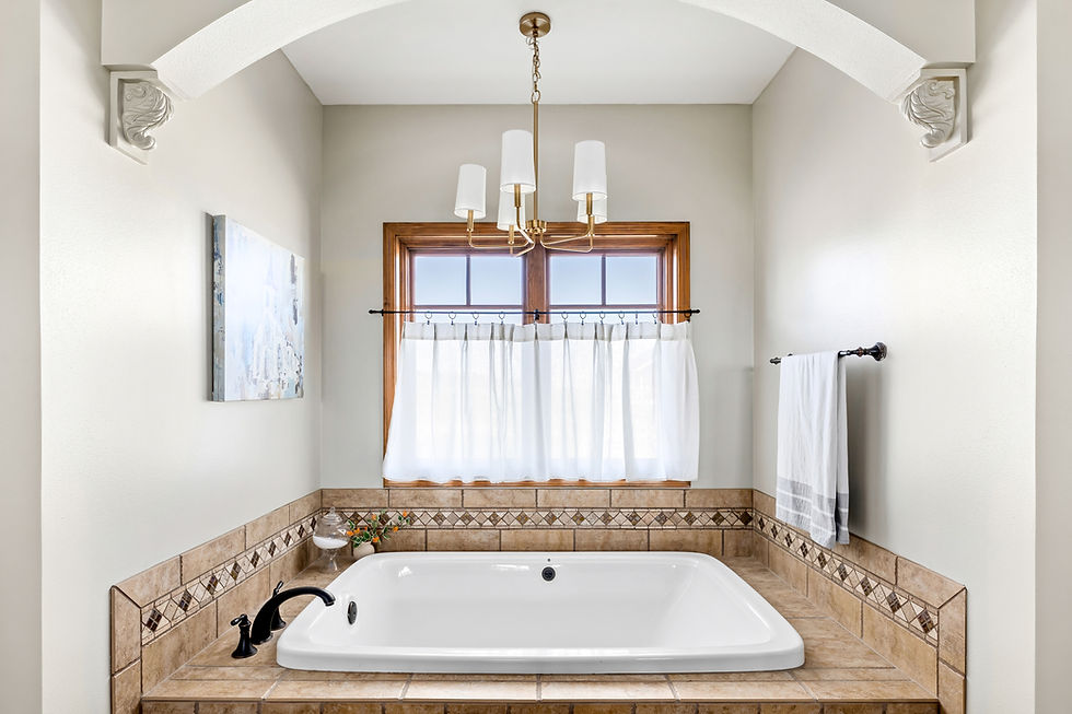 Elegant bathroom nook featuring a deep soaking tub surrounded by tile, beneath a decorative arch and a gold chandelier. Natural light filters in through a curtained window above the tub.