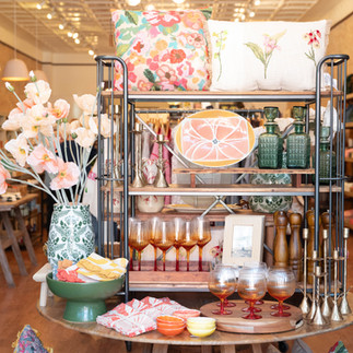 Lifestyle boutique display with colorful pillows, glassware, vases with flowers, and home décor arranged on tiered shelves.