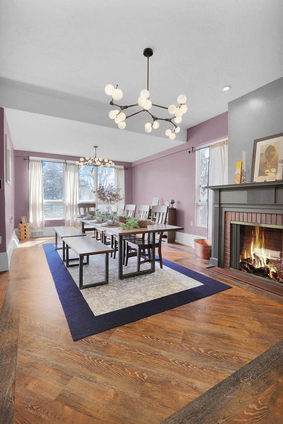 Elegant dining room with a long table, purple walls, two modern chandeliers, and a lit fireplace. Large windows with white curtains. Cozy ambiance.