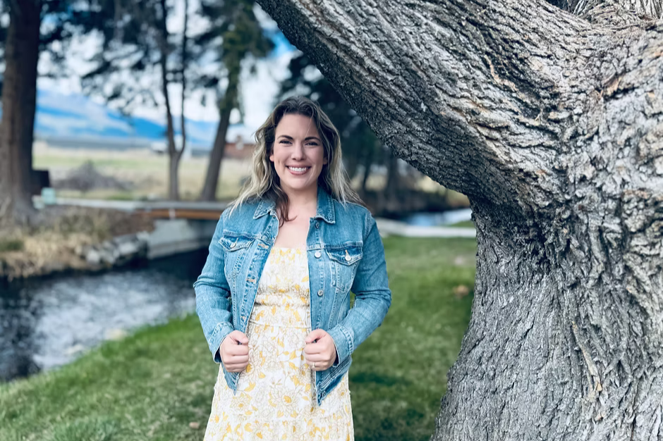 Woman smiling in a denim jacket and yellow floral dress by a large tree and stream, with a blurred forest and mountain in the background.