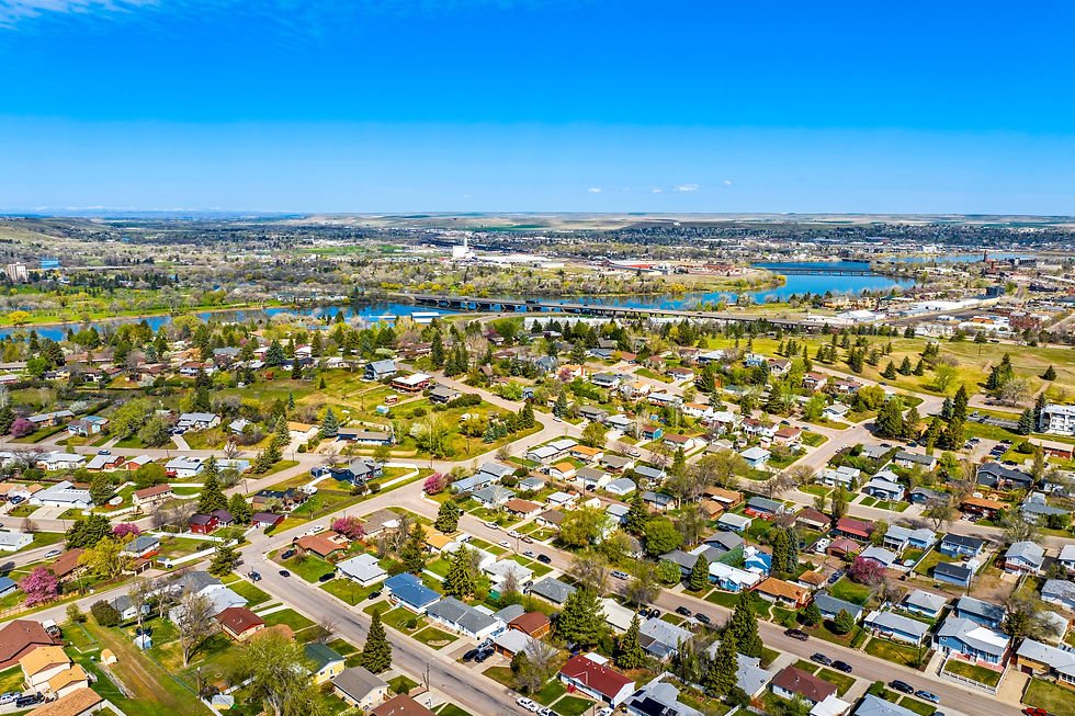Aerial view of a residential neighborhood in Great Falls, Montana, showing a mix of single-family homes, tree-lined streets, and spring foliage. In the distance, the Missouri River winds through the landscape, with bridges connecting different parts of the city. The image captures a bright, clear day with expansive views of the surrounding valley and distant hills under a vivid blue sky.