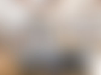 Bright and inviting living room with large picture windows overlooking rolling hills and a river in the distance. The space features wood-beamed ceilings, a stone fireplace with a mounted TV, and two tufted gray sofas arranged around a central table. Natural wood trim and hardwood floors add warmth to the room’s modern rustic charm.