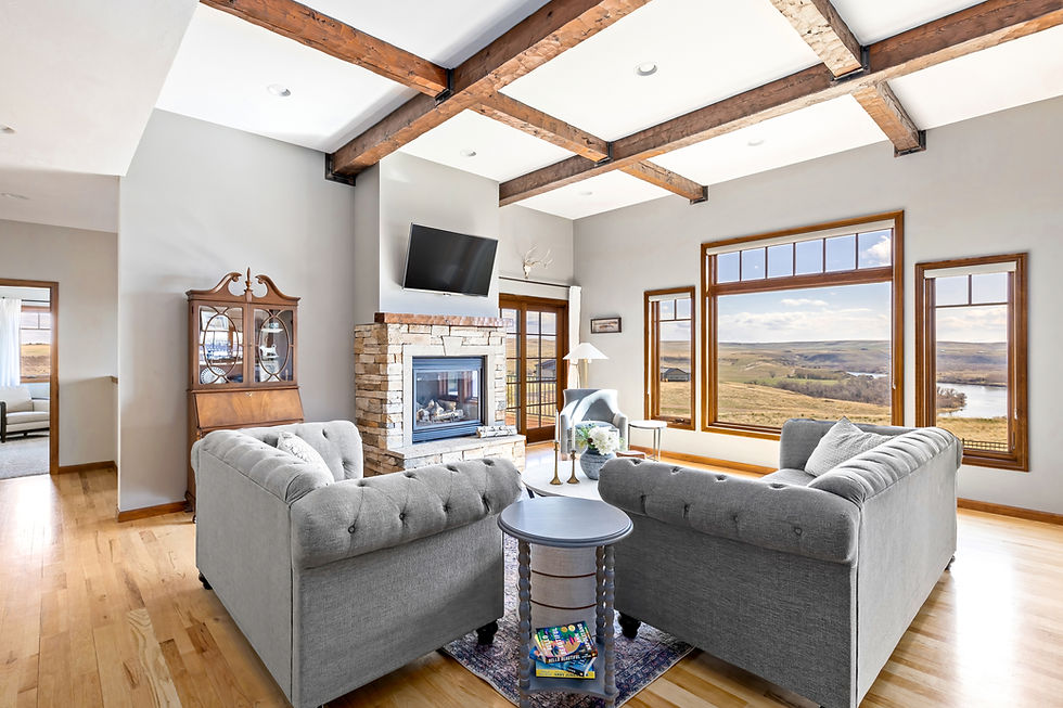 Bright and inviting living room with large picture windows overlooking rolling hills and a river in the distance. The space features wood-beamed ceilings, a stone fireplace with a mounted TV, and two tufted gray sofas arranged around a central table. Natural wood trim and hardwood floors add warmth to the room’s modern rustic charm.