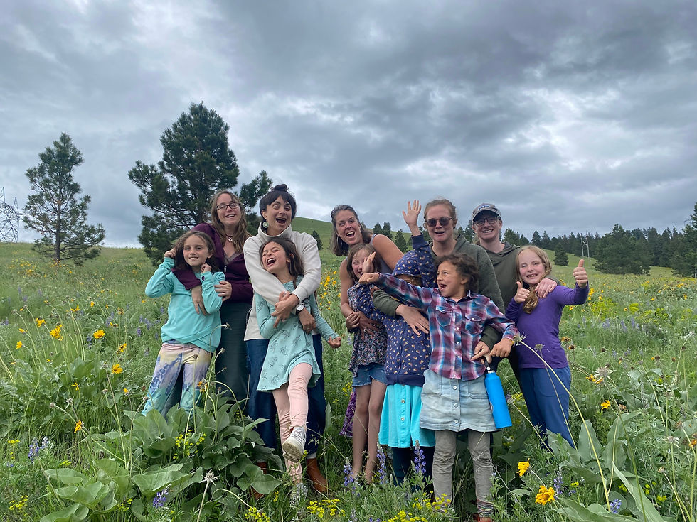 Group of smiling people, including children, posing in a field with wildflowers and trees. Overcast sky, vibrant clothing, joyful mood.