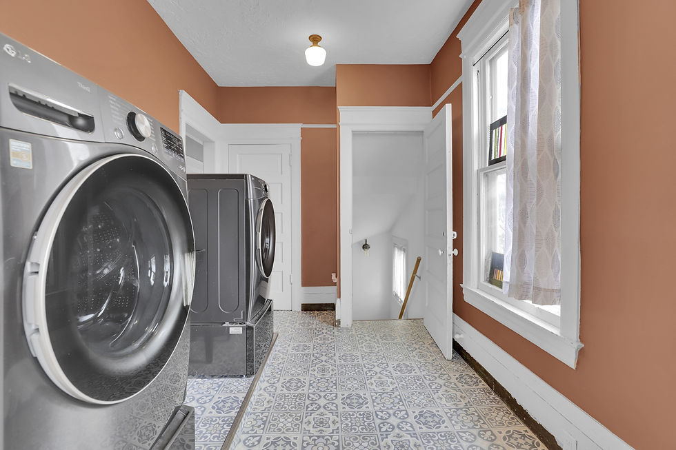 Laundry room with two silver washing machines, patterned tile floor, and brown walls. A window with sheer curtains and a white door.