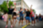 People in festive attire cheer with drinks on a street, surrounded by a crowd near a traffic light. A brick building and trees are visible.