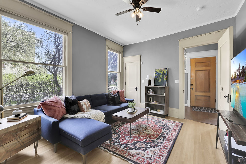 Cozy living room with a blue sectional sofa, colorful pillows, and a patterned rug. A ceiling fan and large windows brighten the space.