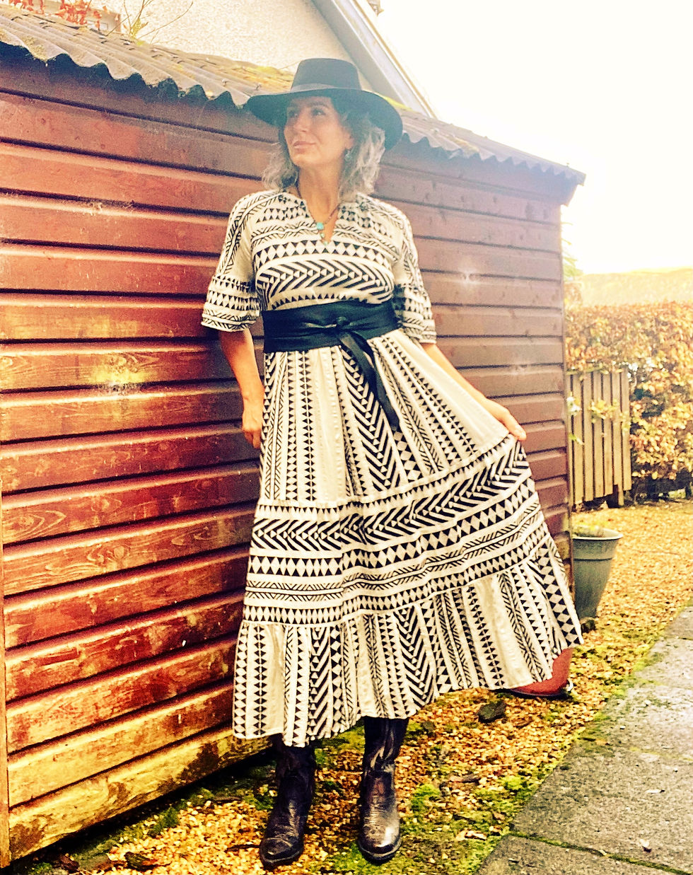 a photo of a woman in a black and white maxi dress styled with Western accessories, standing in front of a wooden outbuilding