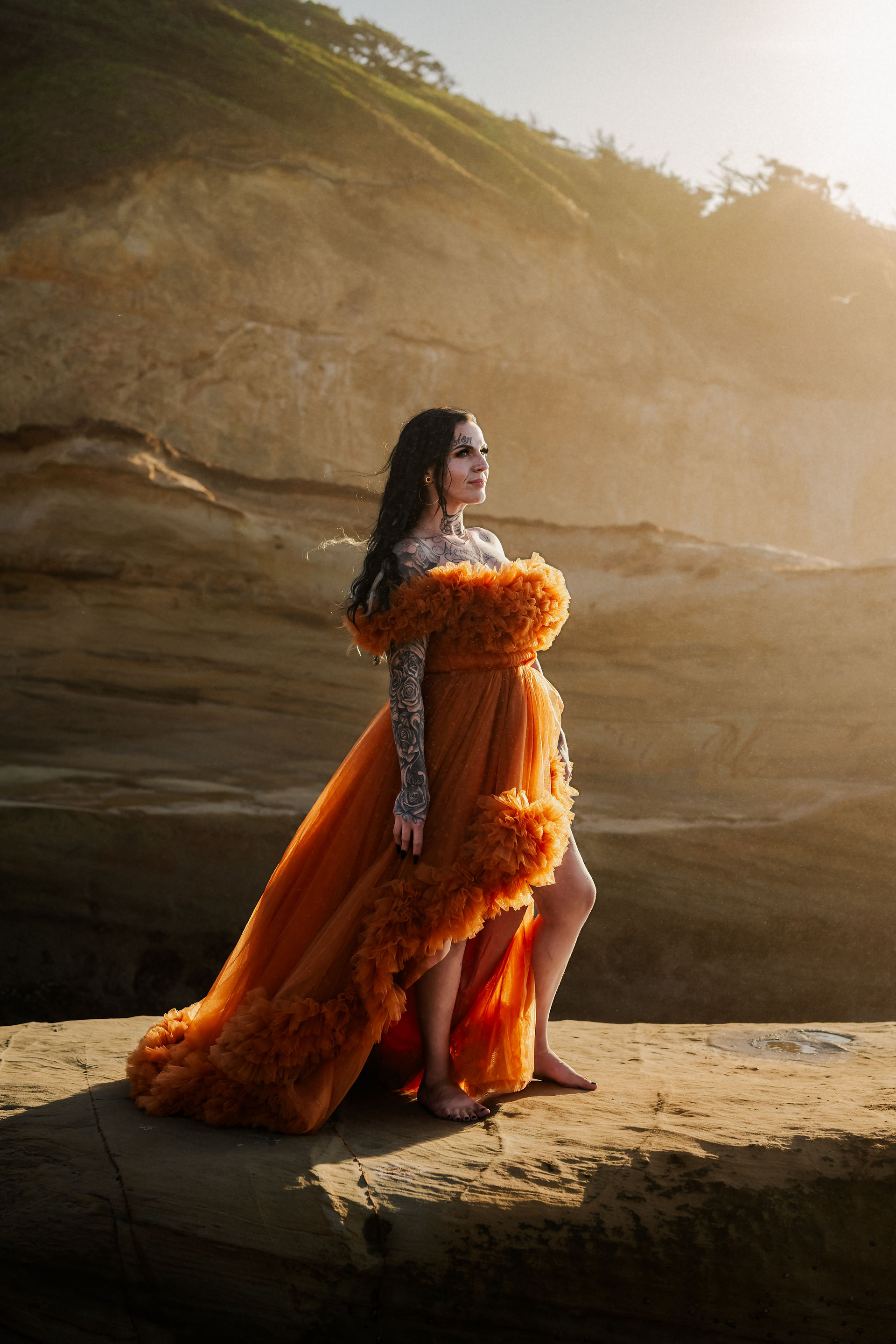 woman in orange dress sands on cliffside during golden hour on the oregon coast at cape kiwanda