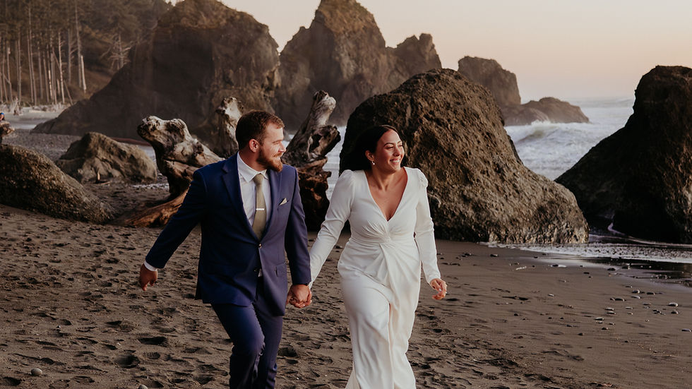 Bride in white gown and groom in blue suit joyfully walk on a rocky beach at sunset, with waves and cliffs in the background.