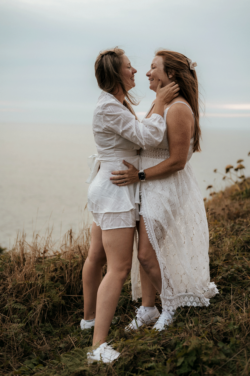 GIF of a lesbian couple on an ocean hillside embrace and kiss. they are wearing all white. it's the day before their elopement. 