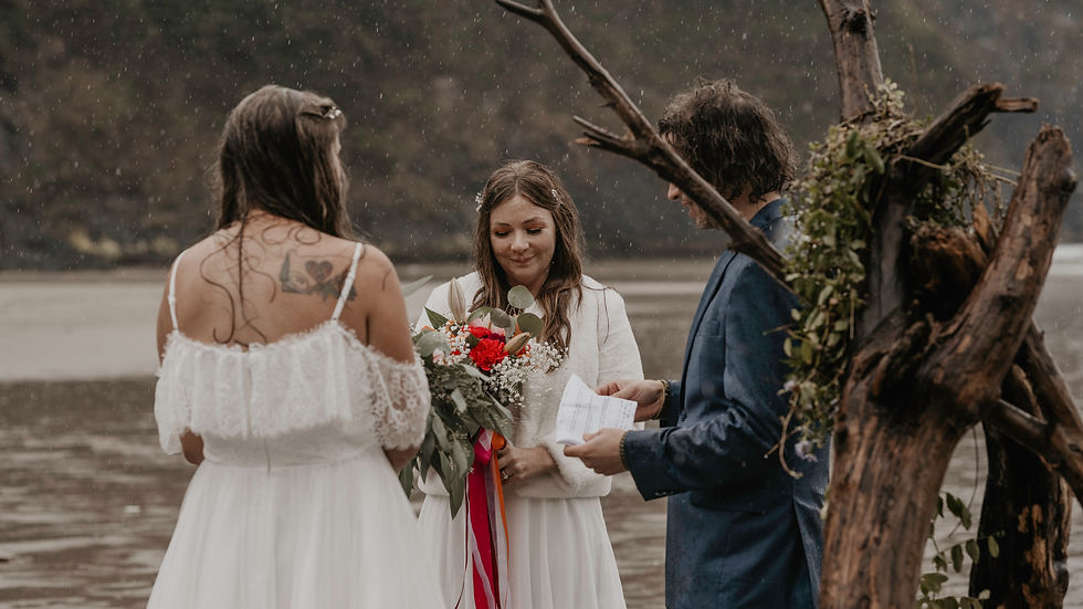 lgbt couple exchange vows on heceta head beach on the oregon coast. it is two brides wearing wedding gowns on the beach. their ceremony is in the rain and a friend is officiating their ceremony.