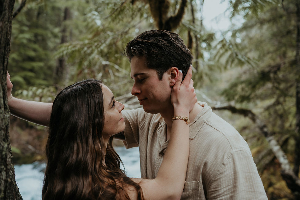 A couple embraces warmly in a lush forest setting, sharing an intimate moment. The background features greenery and a hint of water.