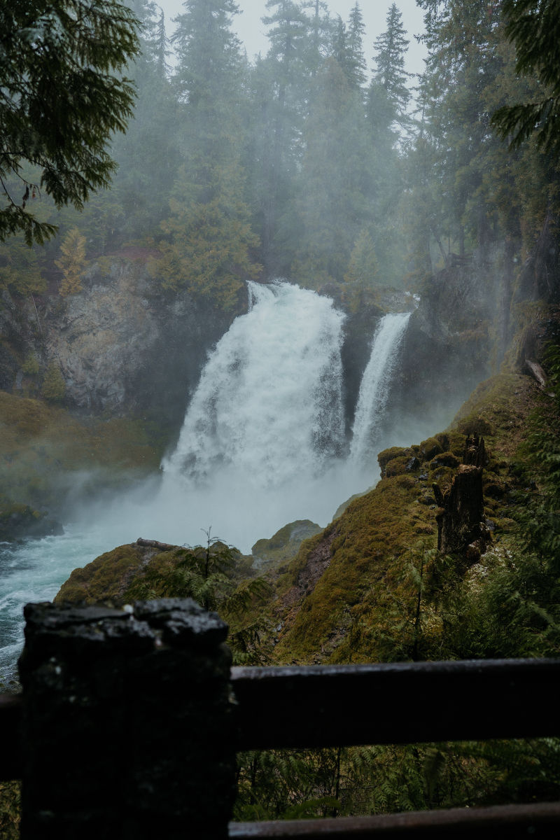 Waterfall cascading down rocks surrounded by lush green forest under a misty sky. Foreground shows a dark wooden fence. Calm, serene mood.