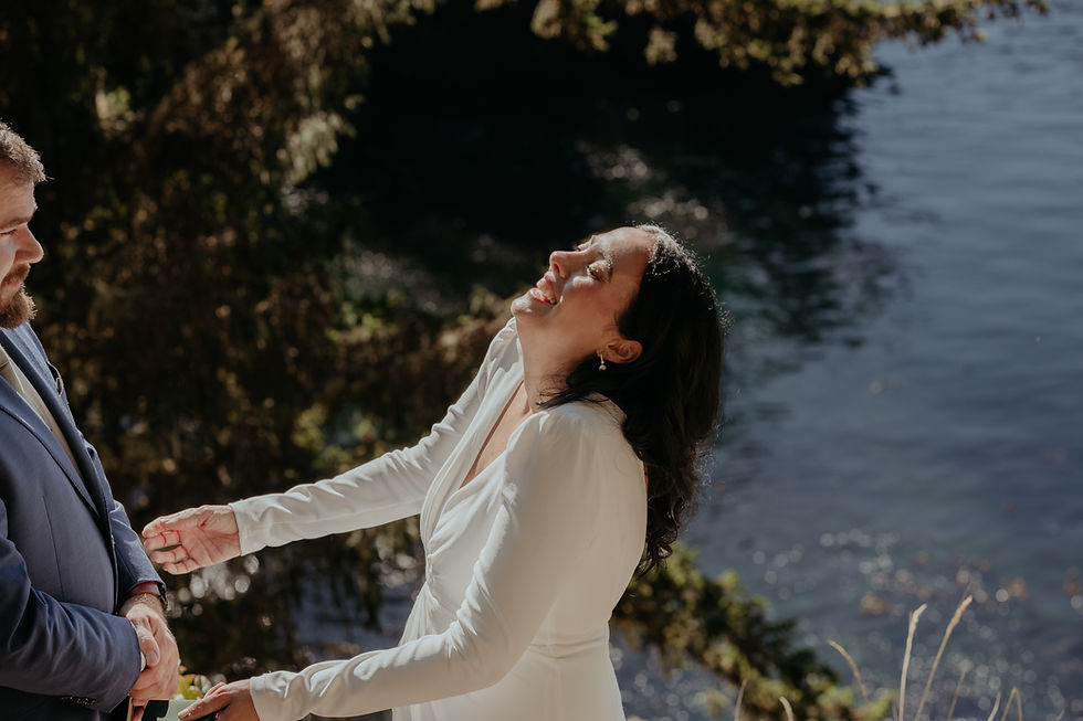 Bride in white laughing joyfully, holding groom’s hands in a blue suit. Background features water and greenery, creating a serene setting.
