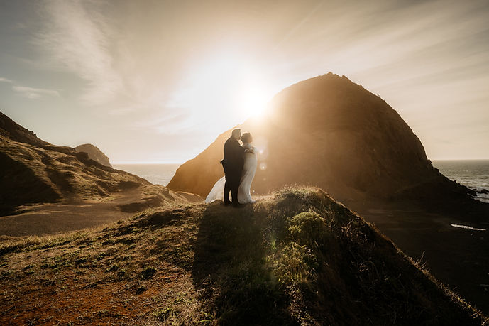an elopement couple stand infront of eachother, both facing the same direction. the groom stands behind the bride and holds out her arms, as if to haver her feel as if she is flying in the swift ocean breeze. they are standing on a hillside ledge that overlooks the pacific ocean in Gold Beach, Oregon. This location on the Oregon Coast is called, "sister's rock". it is an expansive landscape. towering sea rocks form an archipegalo syle landscape, with grassy knolls and a beach down below. it is golden hour. the sunlight pours over the big rock feature and engulfs the couple as they close their eyes and listen to the sound of the ocean waves hitting the rocky shore.