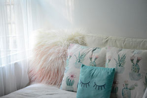 Cozy corner with fluffy pillows, one with cactus print, another in teal with eyelash design; soft light through sheer curtains. Calm mood.
