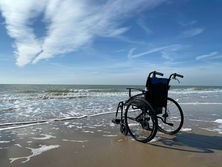 A wheelchair on a sandy beach with waves lapping at the shore, under a bright blue sky with white clouds, evokes solitude and calm.