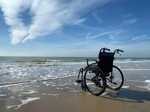 A wheelchair on a sandy beach with waves lapping at the shore, under a bright blue sky with white clouds, evokes solitude and calm.