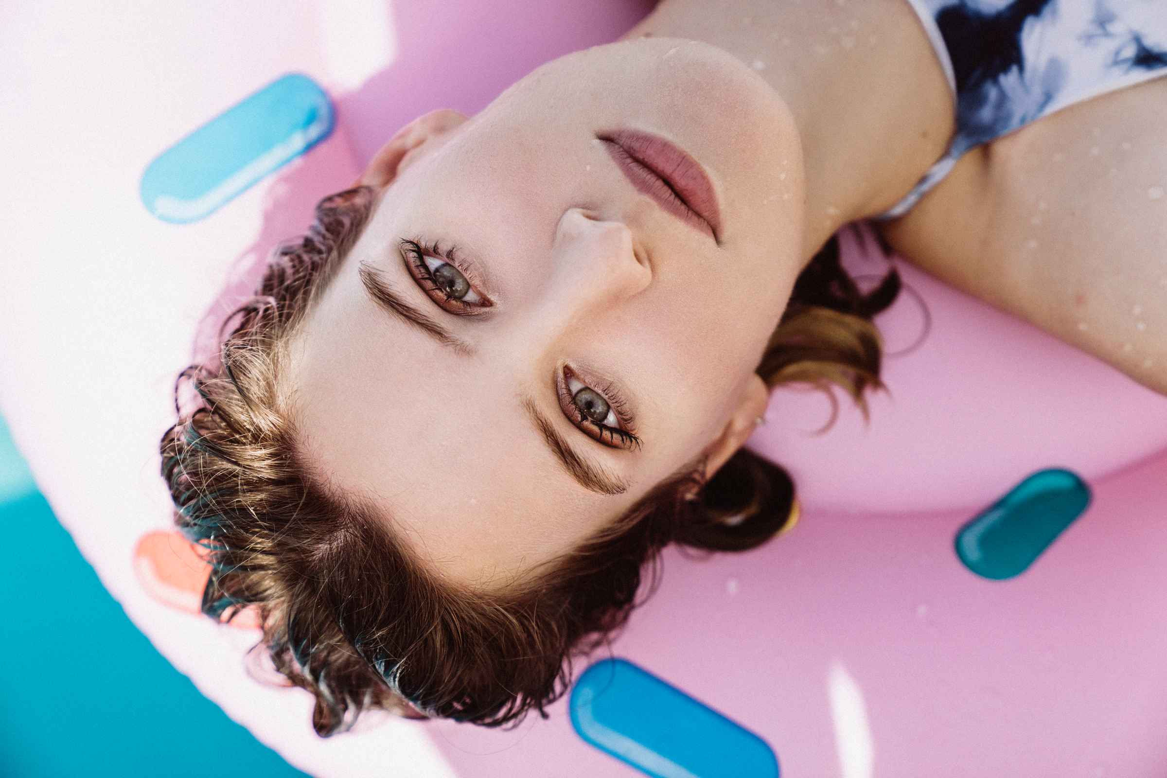 Woman with wet hair lies on a pink inflatable pool float, gazing up. Her expression is calm. The turquoise water is visible in the background, creating a relaxed tone.