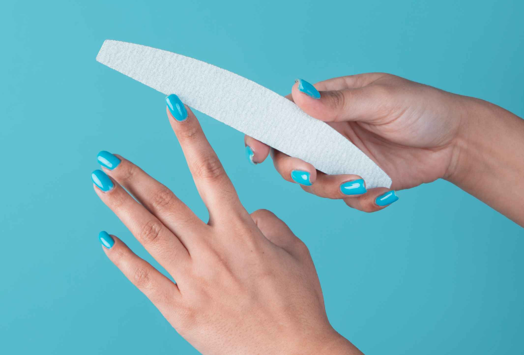 Hands with bright blue nail polish use a white nail file against a light blue background, creating a clean and vibrant self-care scene.