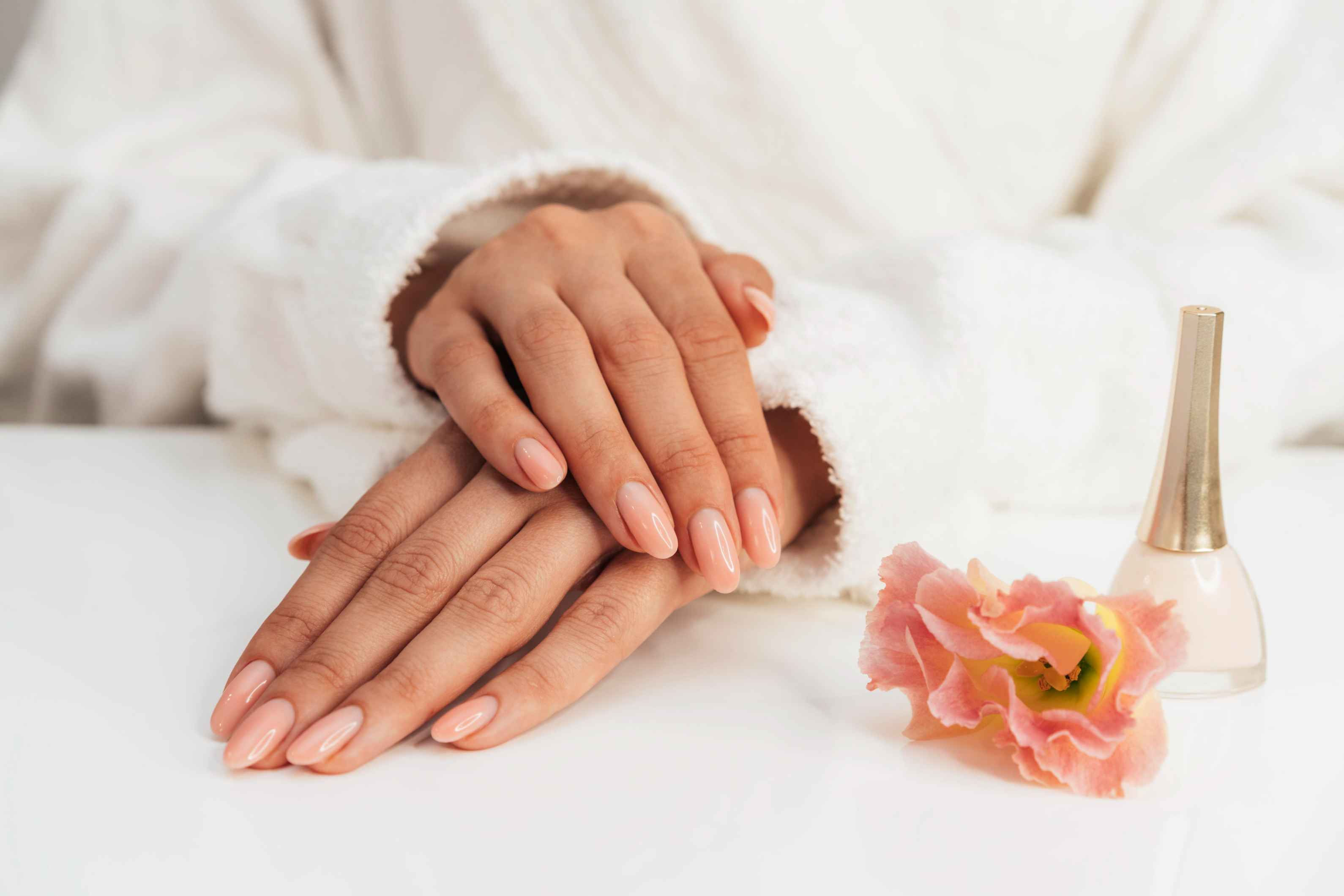 Hands with manicured nails rest on a white surface, wearing a soft robe. Nearby are a pink flower and a bottle of nude nail polish, evoking elegance and tranquility.