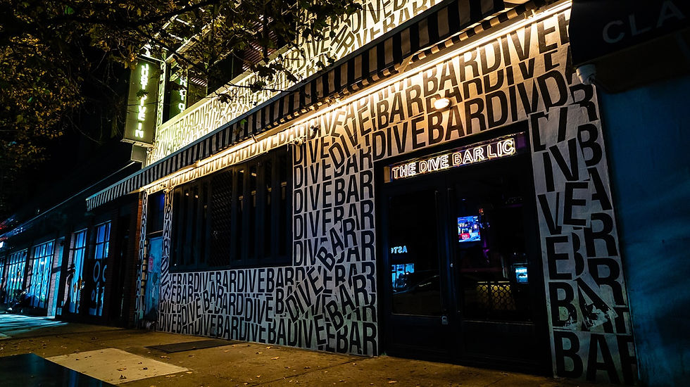 Neon-lit facade of "The Dive Bar", clad in bold black "DIVE BAR" text. Nighttime city street ambiance with dim lighting.