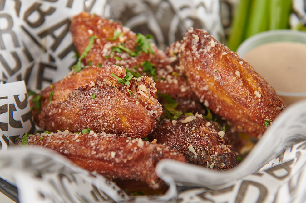 Close-up of crispy fried chicken wings with herbs, resting on printed paper. Dipping sauce and celery sticks in the blurred background.
