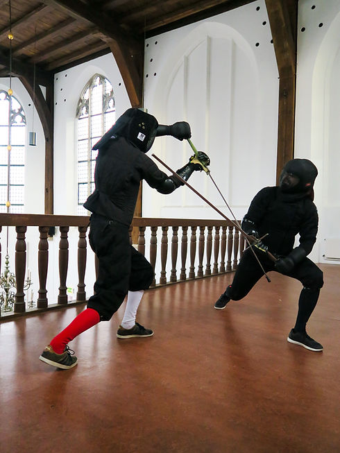 Twee schermers op een balkon in een kapel. De linkerschermer wijst met zijn zwaard naar de grond, terwijl de rechter met zijn kling eraan gebonden is en met de punt naar het gezicht van de linkerschermer wijst.