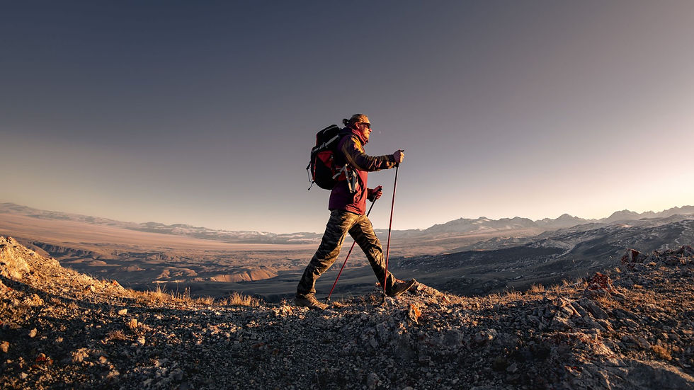 Hiker with poles walks on rocky terrain at sunset, wearing a backpack. Vast mountainous landscape stretches under a clear sky.
