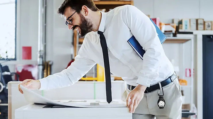 Man in white shirt and tie smiles while examining large papers in a bright office. A blue folder rests in his arm; a watch dangles from his belt.