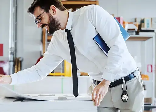 Man in white shirt and tie smiling, examining blueprints in bright office. Holds a blue folder; industrial shelves in background.