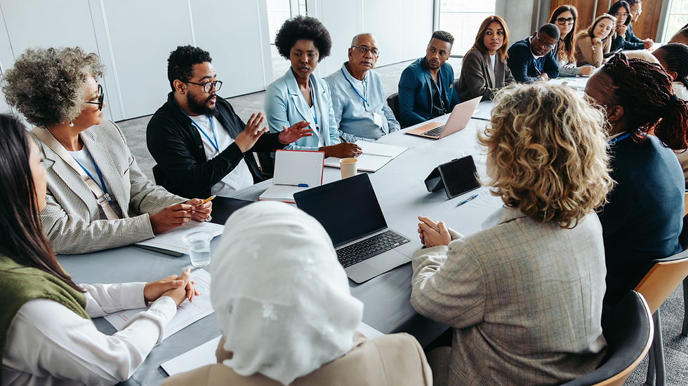 Diverse group in a meeting room, discussing around a table with laptops and notes. Bright setting, serious and focused mood.