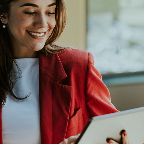 Smiling woman in red blazer holding a tablet, in a bright, blurred office setting. Mood is positive and engaged.