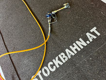 Nahaufnahme Stockbahn-Oberfläche mit gelbem Schlauch und Logo STOCKBAHN.AT – rutschhemmende Beschichtung