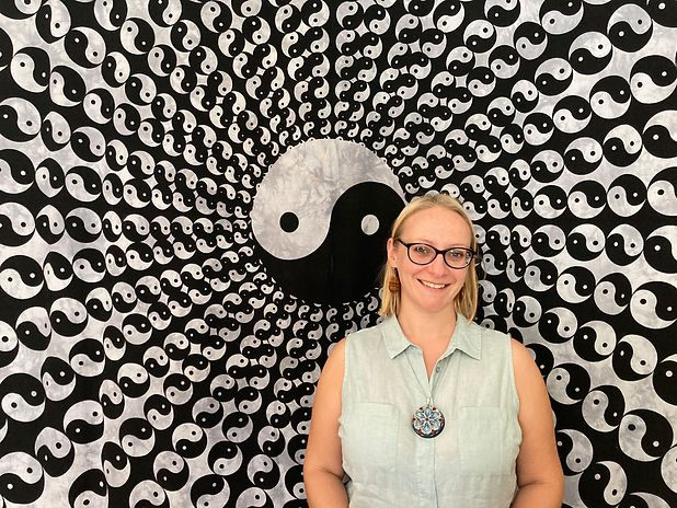 Headshot of female with shoulderlength blonde hair wearing seeing glasses standing in front of an image of a ying and yang symbol