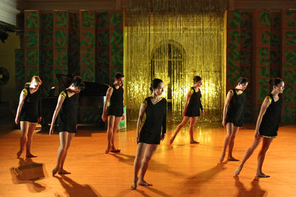 A line of dancers in black tunics and bare feet tilt in unison on a golden-lit stage, framed by glittering tinsel and patterned panels.A line of dancers in black tunics and bare feet tilt in unison on a golden and orange stage, framed by glittering tinsel and patterned panels.