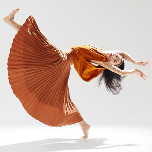 A dancer in a flowing orange top and wide pleated skirt arcs midair, body tilted sideways with outstretched arms against a bright studio backdrop.