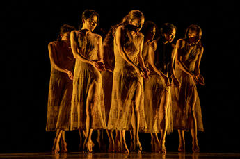 A tight line of dancers in identical golden dresses stands barefoot, hands softly cupped, glowing under warm stage light against a dark background.