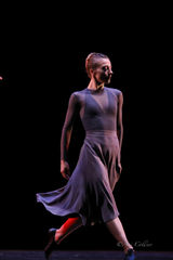 A solo dancer in a flowing purpule dress strides across a dark stage, body angled and gaze turned aside, captured mid-motion.